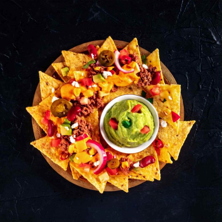 A circular plate filled with a homemade nachos recipe that has tortilla chips with ground beef, tomatoes, onions, jalapenos, cheese, kidney beans, avocado and corn on top, with a small ramekin of guacamole on top garnished with cilantro and more diced tomatoes.