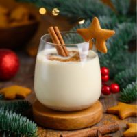 A glass filled with a generous serving of homemade non alcoholic egg nog on a wooden coaster, garnished with a cinnamon stick and ginger bread cookie on the glass, surrounded by pine boughs, red Christmas ornaments, more gingerbread cookies, cinnamon sticks and star anise with a bokeh of lights in the background of the image.