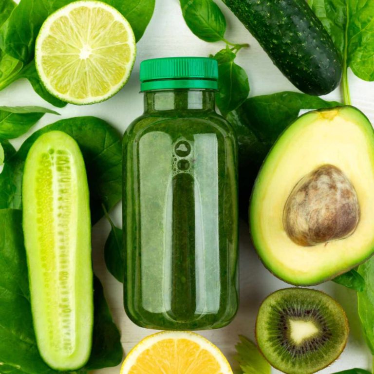 A close up shot of a bottle of cold pressed green juice containing an easy green juice recipe, resting atop various greens including spinach, limes, kiwi, avocados and cucumbers.