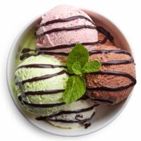 A white bowl with four scoops of ice cream in it in varying colors (pink, green, brown and white) with a chocolate sauce drizzled on top, and garnished with a sprig of mint to celebrate Ice Cream For Breakfast Day.
