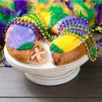 A Mardi Gras king cake on a cake stand with Mardi Gras beads draped on top of it, and a plastic king cake baby sitting in the space where a piece of cake was removed, with purple, green and gold yellow in the background.