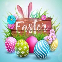 A wooden sign that reads Happy Easter surrounded by decorated easter eggs, with flowers and grass around the sing and a pair of bunny ears peeking up behind the sign.