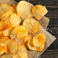 A pile of homemade potato chips with sea salt piled on a piece of parchment paper on a wooden background.