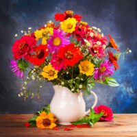 A white porcelain pitcher being used as a vase sitting on a wooden table against a blue background with various wildflowers in red, yellow, purple and white inside of it.