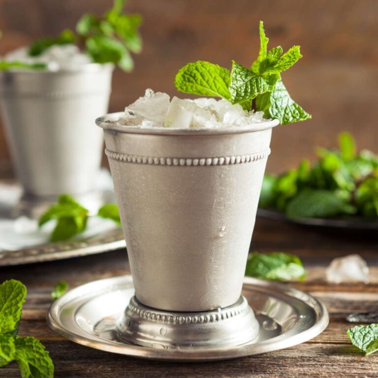 A mint julep garnished with mint on a silver saucer, in a traditional mint julep silver cup, with another mint julep and fresh mint in the background.