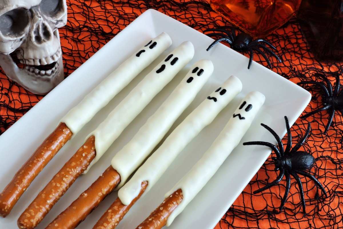 A white plate with five ghost pretzel rods with melted white chocolate and a ghost face drawn on them, on an orange tablecloth with a black spiderweb on top of it, and plastic spiders around the plate of ghost pretzels.
