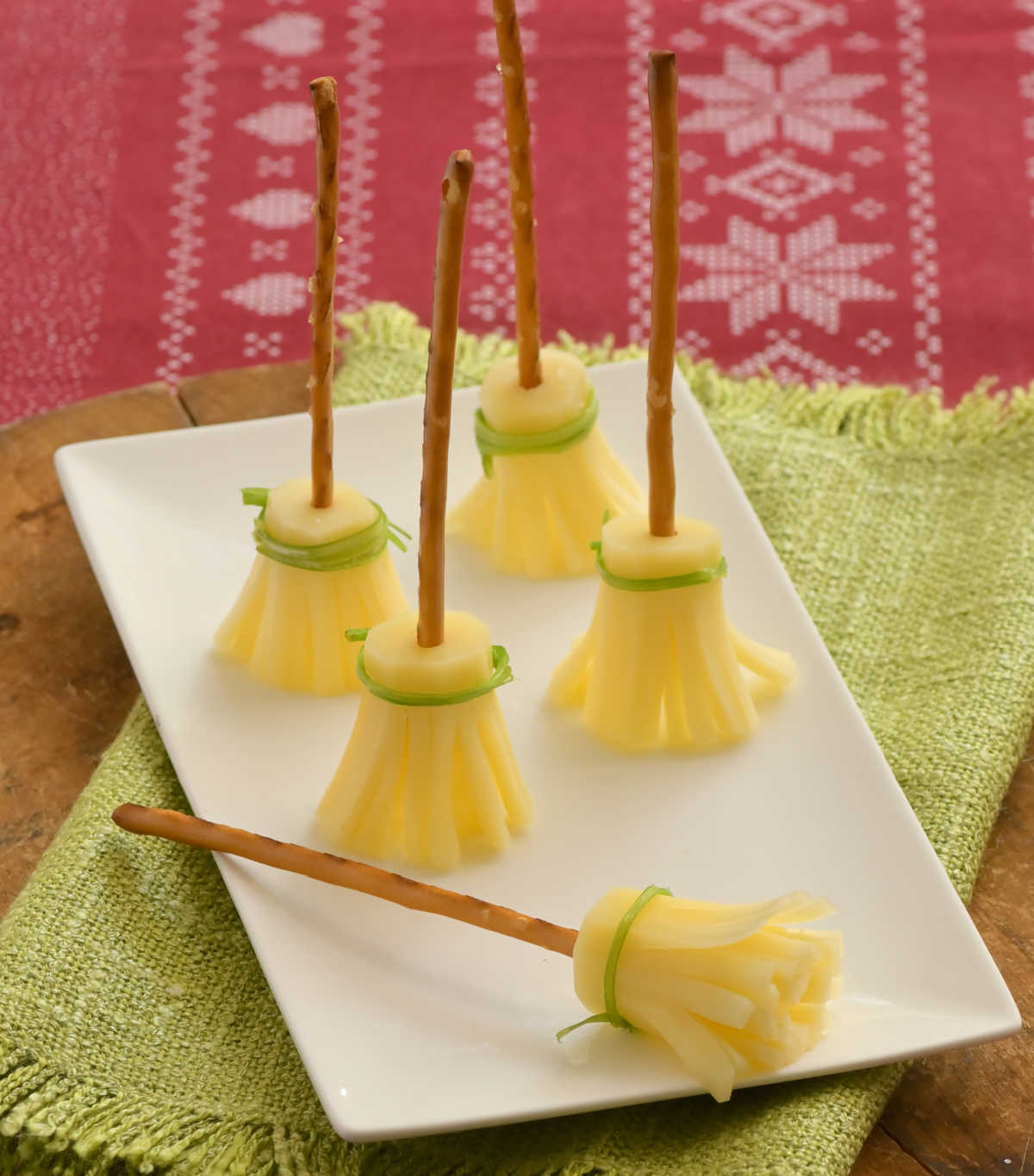 A white plate on a green napkin with 5 DIY witches broomstick treats made out of pretzels, string cheese and chives with four of the string cheese broomstick standing upright, and one laying on its side..