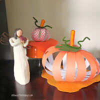 Two DIY paper pumpkins displayed on a mahogany wood table beside an angel figure.