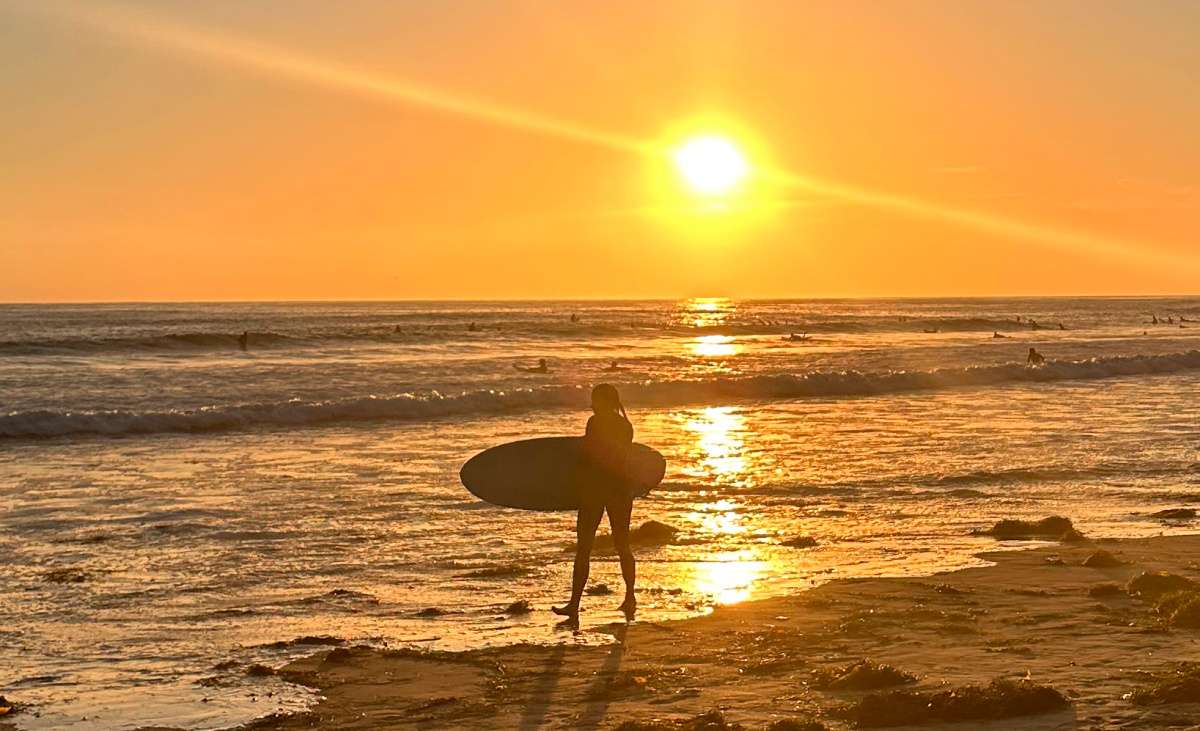 A woman walking on the beach, holding a surf board while the sun is setting behind her during a summer day.