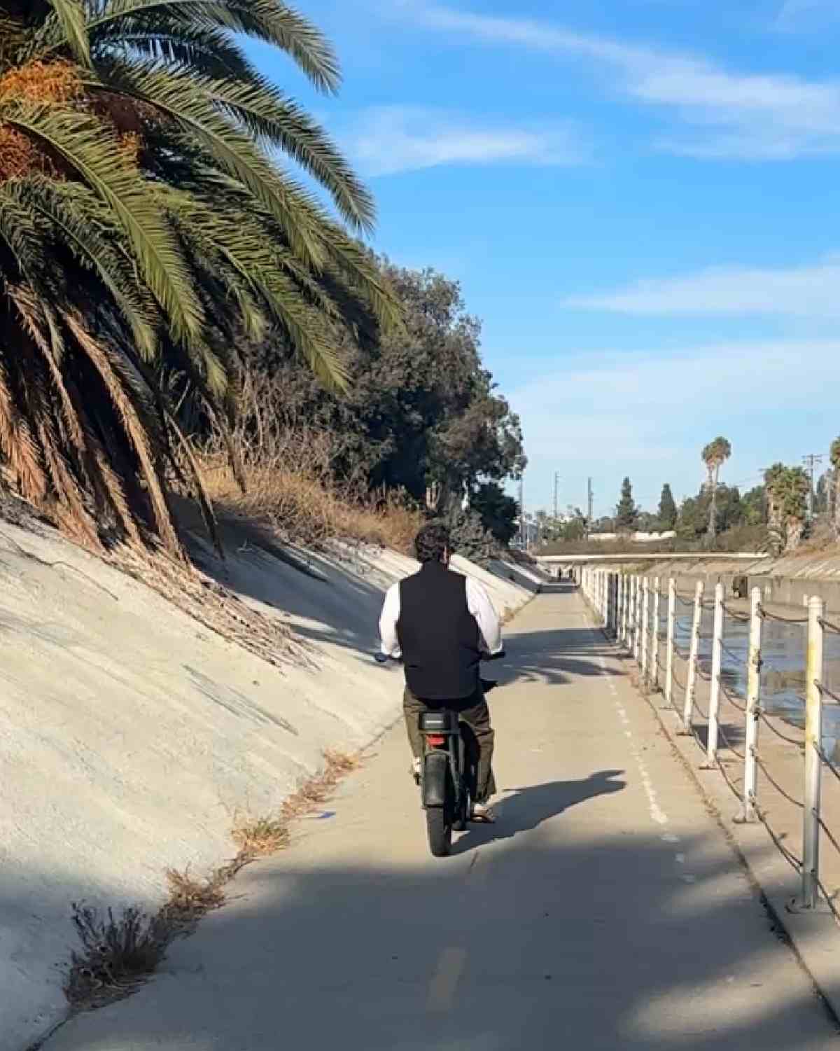 A man riding an electric bike on the Ballona Creek Bike Path with palm trees to his left and the creek to his right.