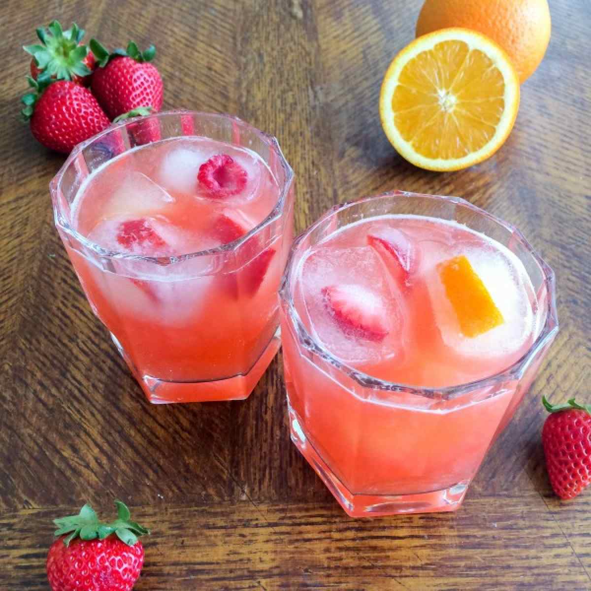 Two glasses filled with tropical mocktails for summer, with fruit ice cubes in the glasses, and fresh strawberries and oranges arranged on the table around the glasses.